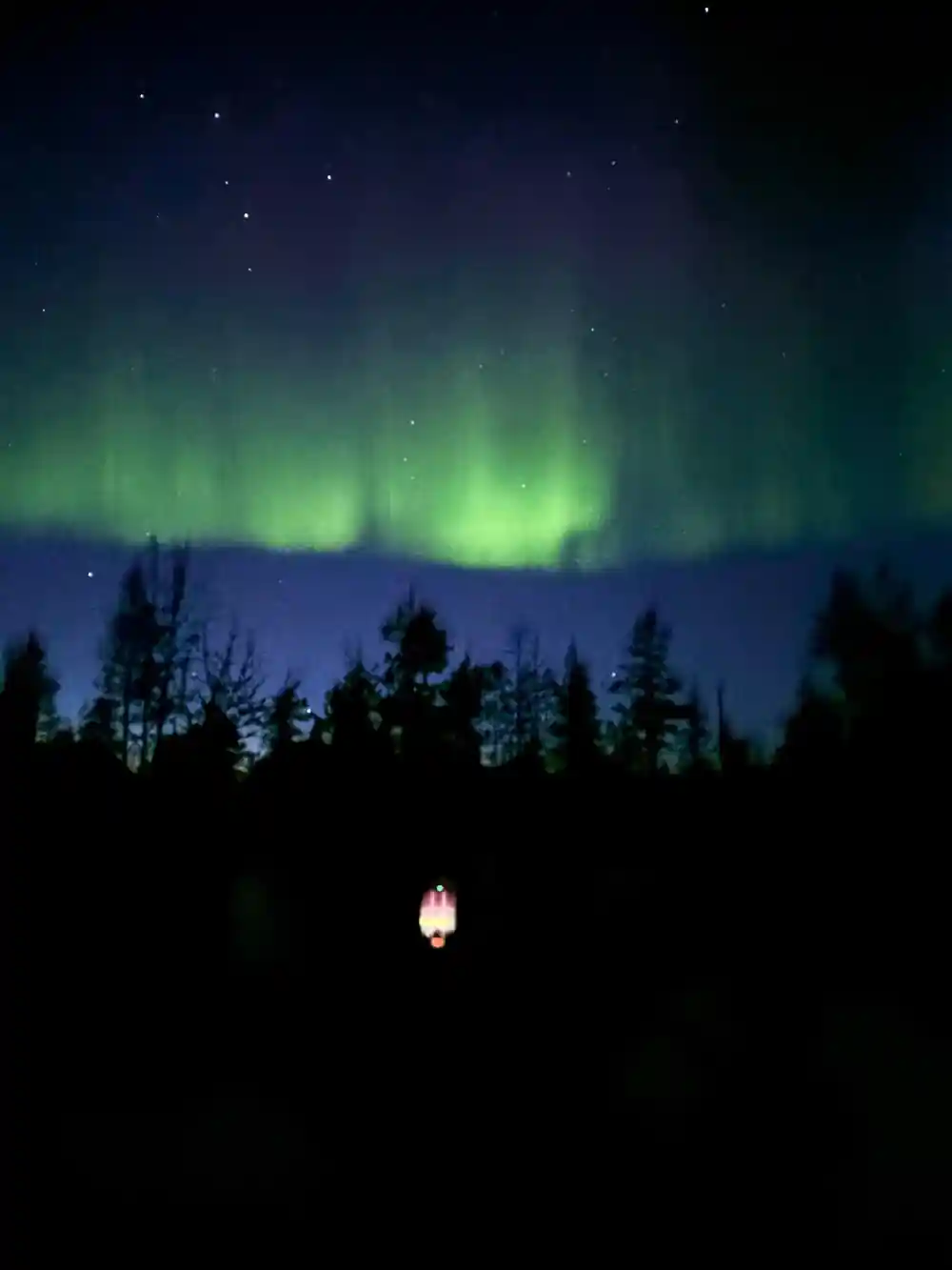 Aurora borealis above forest with single lit cabin below