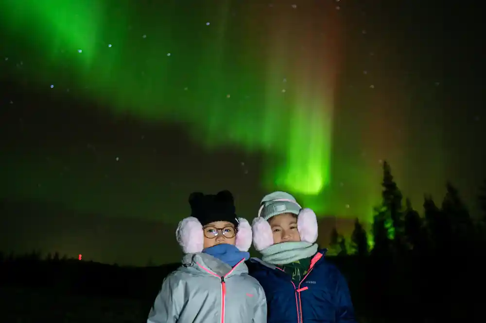 Two children wearing winter hats under green aurora